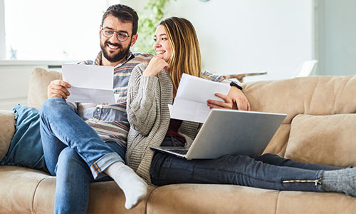 Man and woman comfy on couch while man holds document in hand and woman rests laptop on legs.