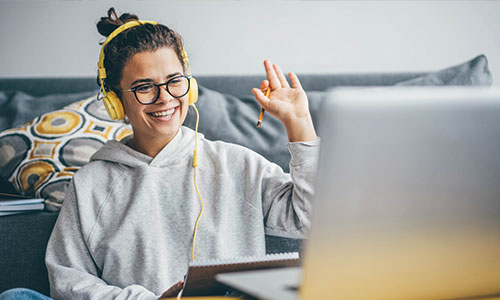 Young woman with headphones on waving to computer screen while sitting on floor in front of couch.