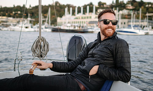 Relaxed man with sunglasses sitting on sailboat while looking out at ocean, with other boats in background.