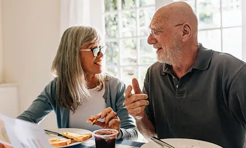 Older man and woman sitting together for breakfast and looking at one another to discuss document in front of them.