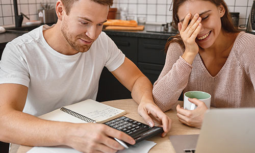 Man and woman sitting together at kitchen table looking at computer screen and using calculator.