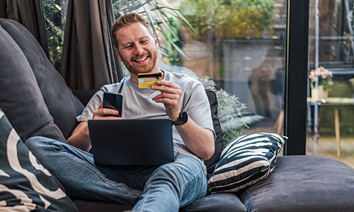 Man relaxing on couch with laptop in lap and phone and credit card in hand.