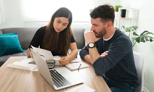 Man and woman sitting at table looking over documents with laptop, notebook and calculator in front of them.