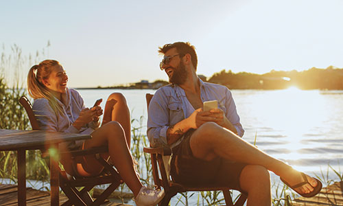 Man and woman relaxing in chairs next to waterfront with sun gleaming behind them.