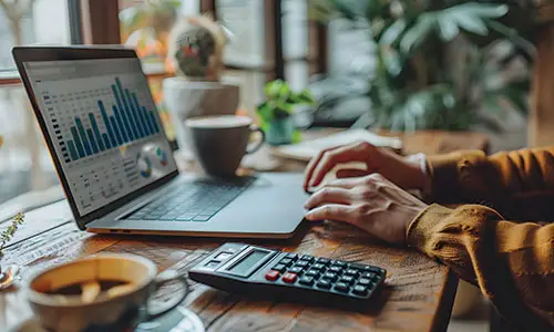 Person working at high top desk with bar charts on laptop screen in front of the them, surrounded by calculator, coffee cup and indoor plants in background.