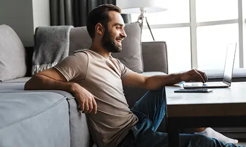 Man sitting on floor with back against couch using laptop on surface in front of him.