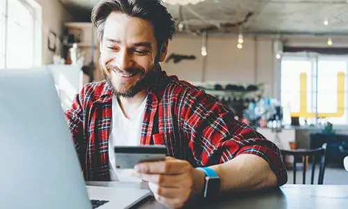 Happy guy smiling at screen while using laptop and holding credit card in hand.
