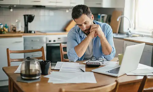Man sitting at kitchen table deep in thought over documents resting on surface with laptop, glasses, and carafe of coffee in foreground.