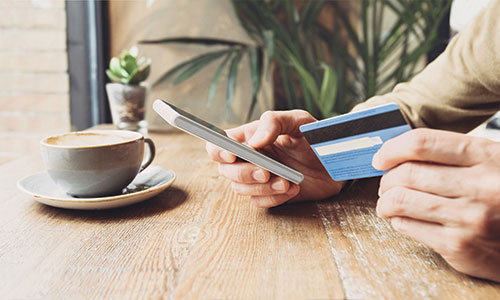 Man sitting at high cafe table with cup of coffee in the foreground, holding phone in one hand and credit card in the other.