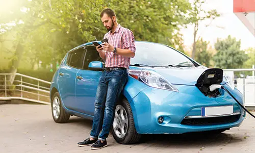 Man resting against side of electric car to use cell phone while car charges.