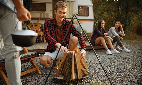 Young man building fire while young women chat in background next to RV.