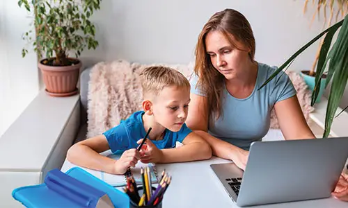 Mother helping son with homework at table with laptop and notebook in front of them.