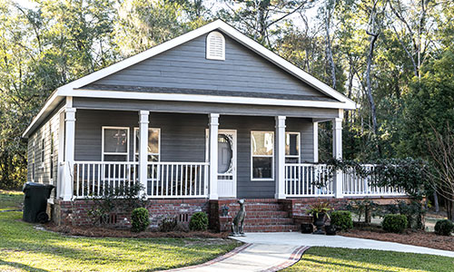 Manufactured home sitting on land with green grass surrounded by trees and sidewalk leading to fron door.