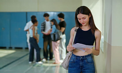 A young woman leans against a wall, reading a paper with a thoughtful expression. In the background, a group of students talks near lockers.