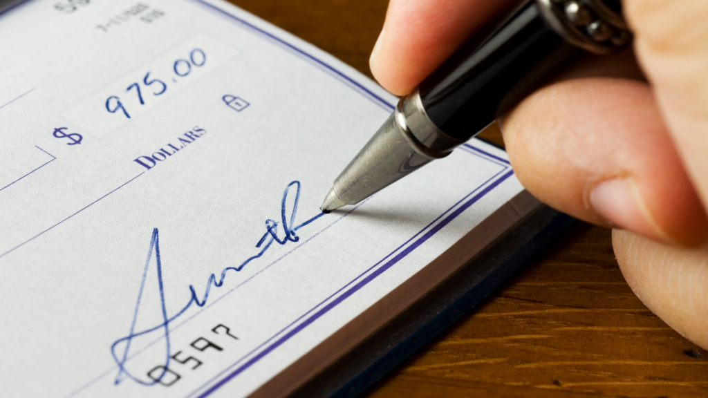 A hand holding a pen signs a check for $975.00. The check is on a wooden table, showing clear, neat handwriting and a formal, professional tone.
